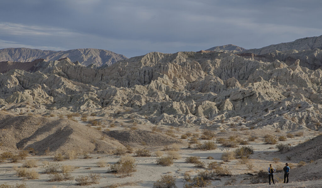 Rugged desert landscape with craggy, eroded hills under a cloudy sky. Two people with backpacks stand at the bottom right, admiring the view.