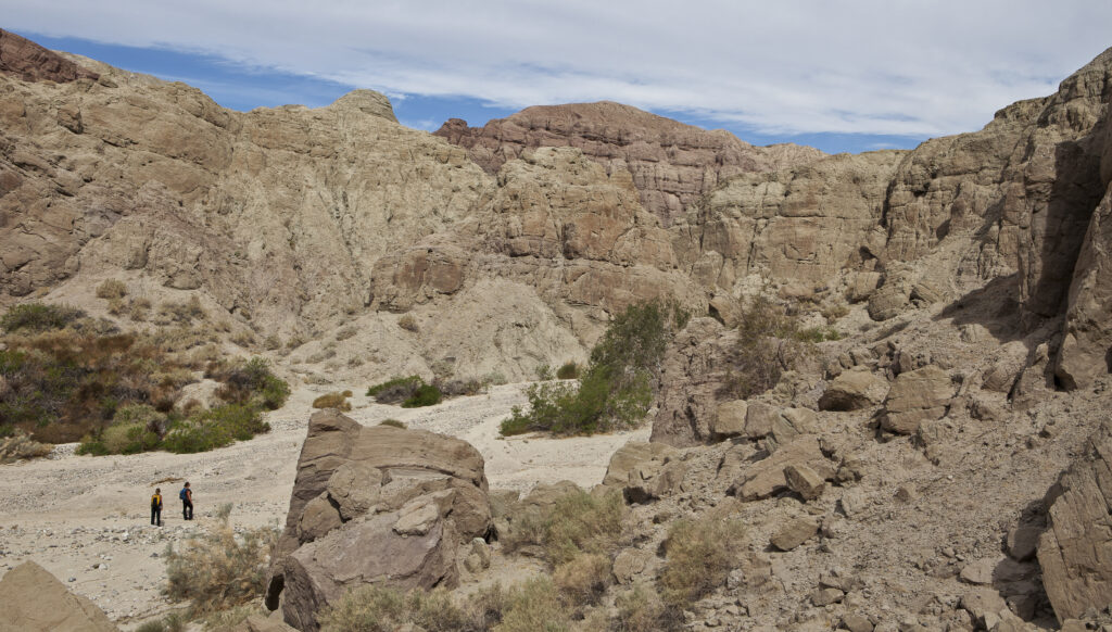 Two hikers walk through a desert canyon with rugged, tan rock formations. Sparse green vegetation contrasts with the arid landscape under a partly cloudy sky.