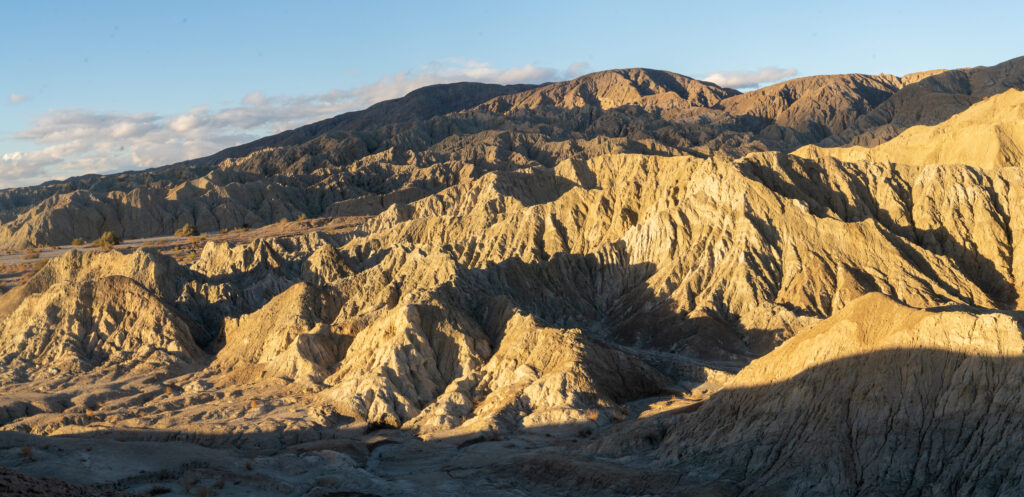 Rugged desert landscape at sunset, featuring layers of sunlit, rocky hills casting dramatic shadows. The sky is clear with a few scattered clouds.