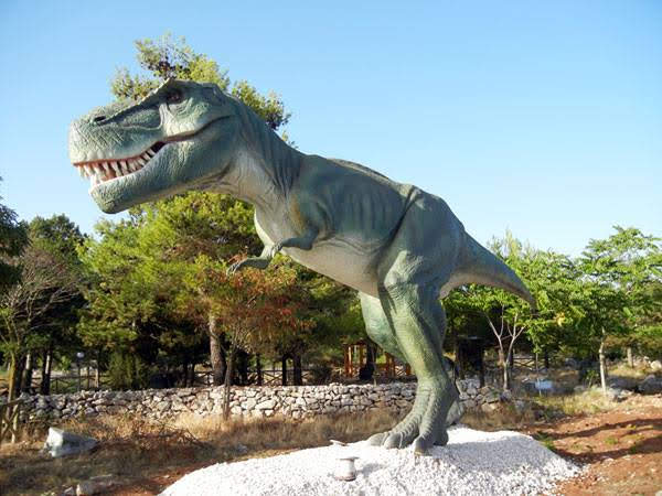 A life-sized Tyrannosaurus rex sculpture stands outdoors on gravel, surrounded by trees. The dinosaur's mouth is open, displaying sharp teeth.