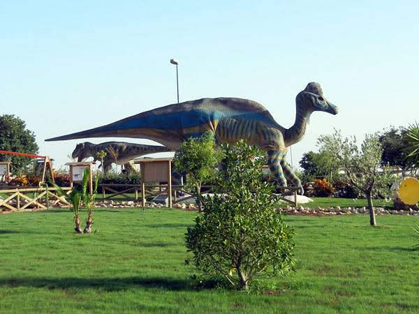 Life-sized dinosaur statues in a park with green grass and trees, under a clear blue sky. The scene is calm and inviting with a playful tone.