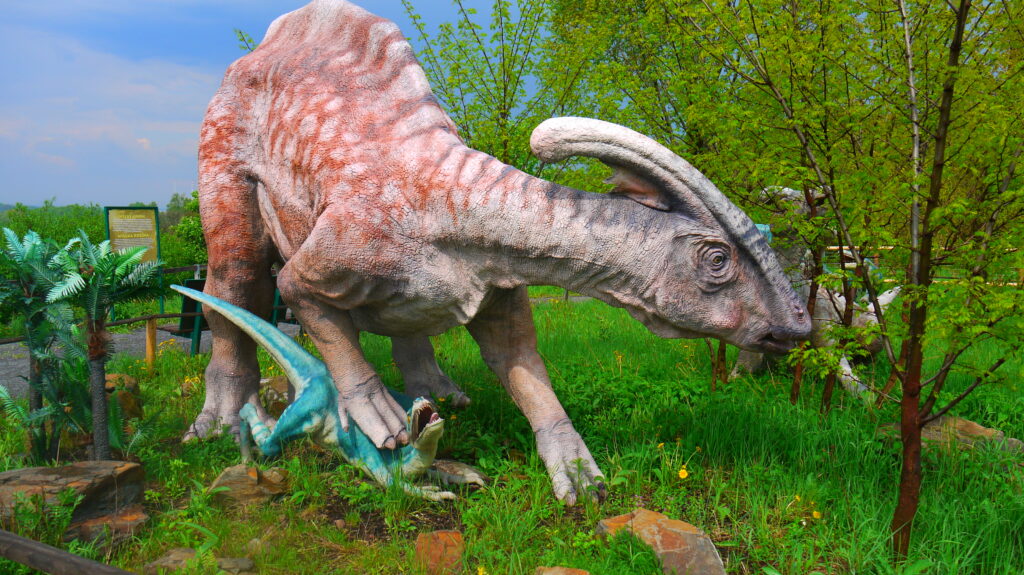 Life-sized statues of dinosaurs in a park setting. The large dinosaur is grazing near trees, while a smaller one stands nearby, surrounded by greenery.