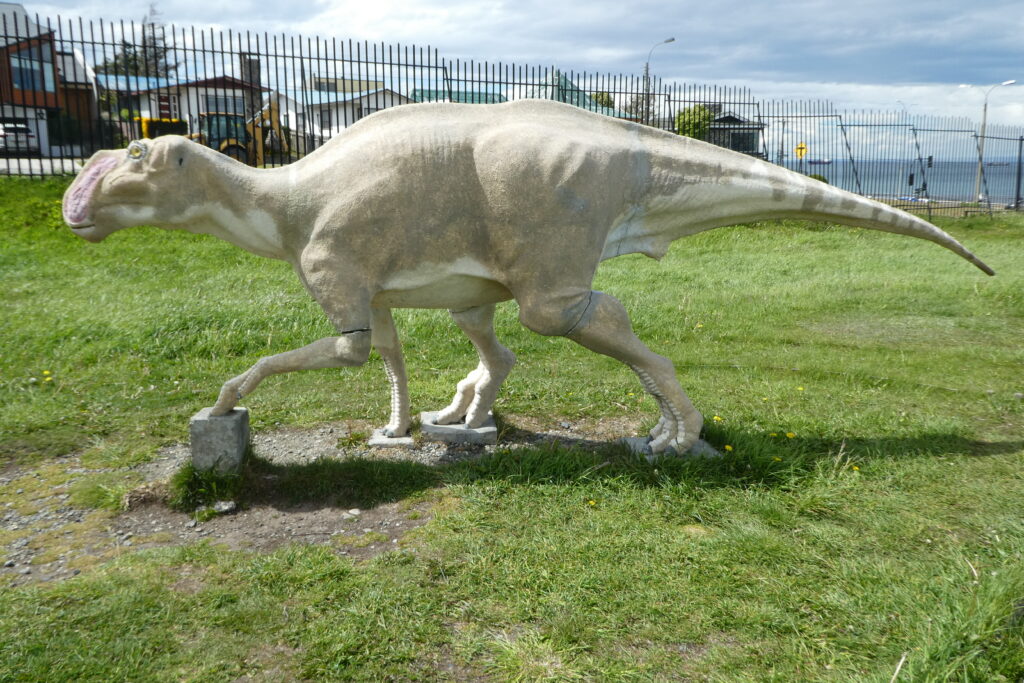A dinosaur sculpture stands on a grassy field with a wire fence and houses in the background. The dinosaur is beige, with a long tail and a large jaw.