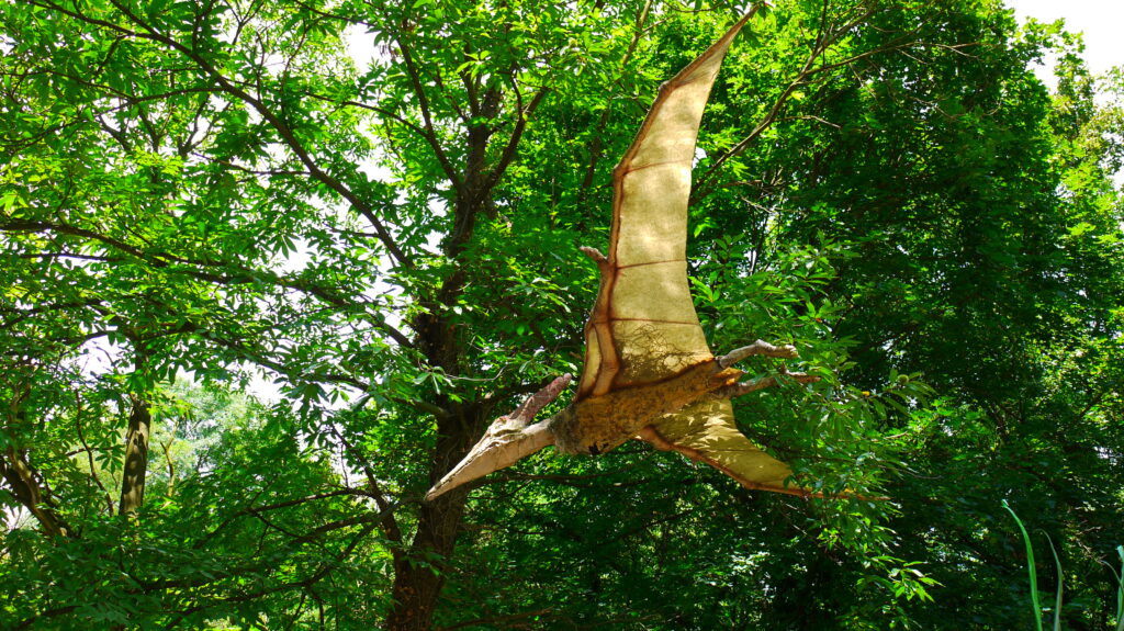 A realistic pteranodon model is suspended among lush green foliage, conveying a prehistoric theme. Sunlight filters through the leaves, creating a vibrant, natural backdrop.