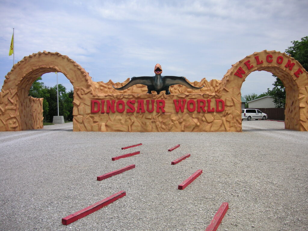 Entrance to Dinosaur World with large gates featuring a dinosaur over the words “Dinosaur World,” flanked by archways and a gravel path.