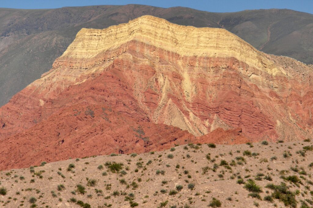 Rocky landscape with layered earth tones, featuring striking red and beige strata against a backdrop of green hills under a clear blue sky.