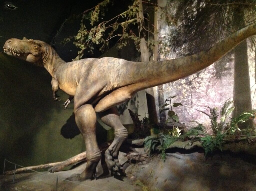 Life-sized dinosaur model on display in a museum, surrounded by artificial plants. The dim lighting and greenery create a prehistoric atmosphere.