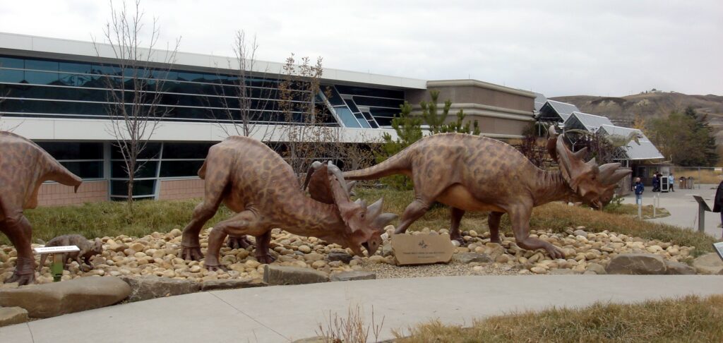 Dinosaur statues of a Triceratops family on rocky terrain outside a modern building. Overcast sky and atmospheric setting suggest a museum exhibit.