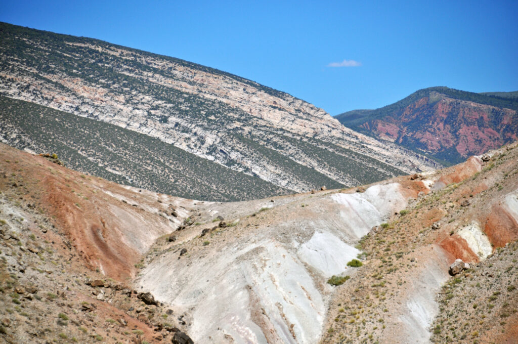 Barren, layered hills with shades of red, white, and brown form rolling slopes under a clear blue sky, evoking a sense of desolation and vastness.