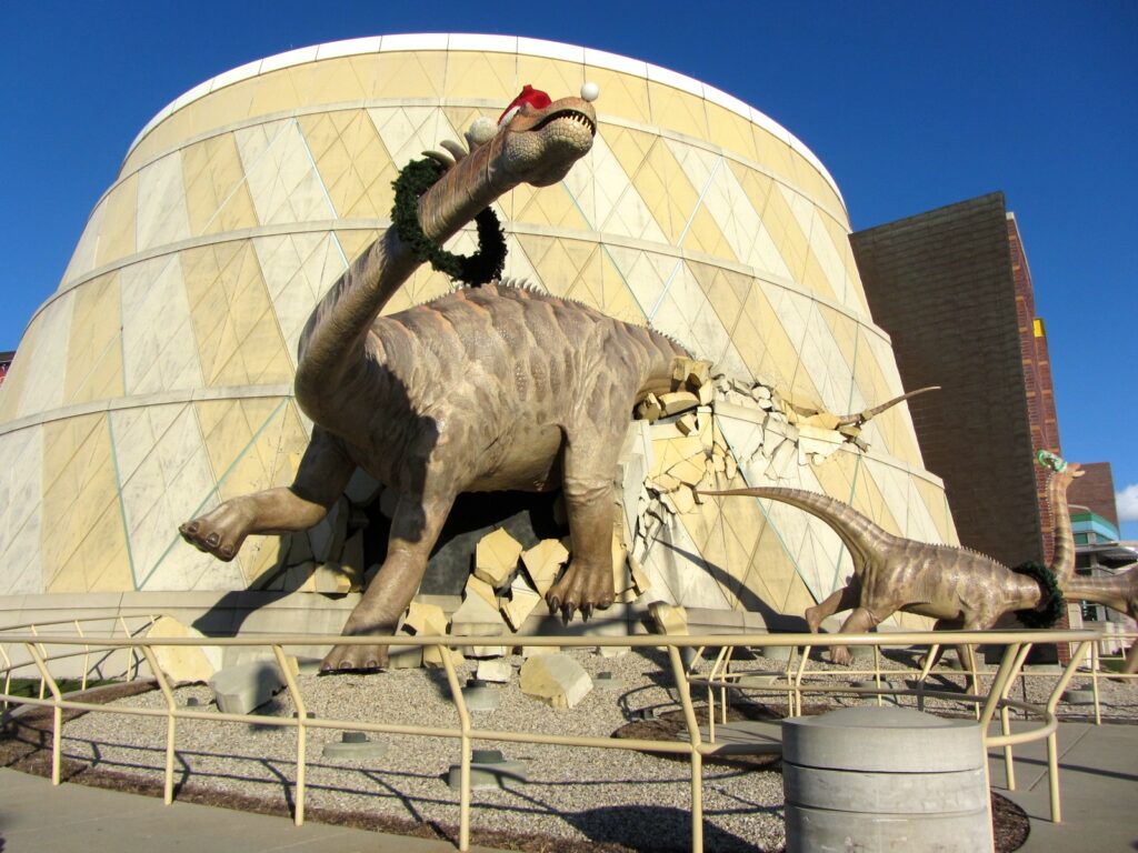Life-sized dinosaur sculptures burst through a building facade. The largest dinosaur wears a Santa hat, creating a playful and lively scene.