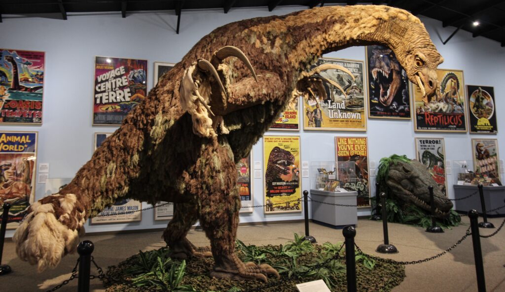 A museum exhibit displays a lifelike dinosaur model with a long neck and clawed hands, surrounded by vintage dinosaur movie posters on the walls.