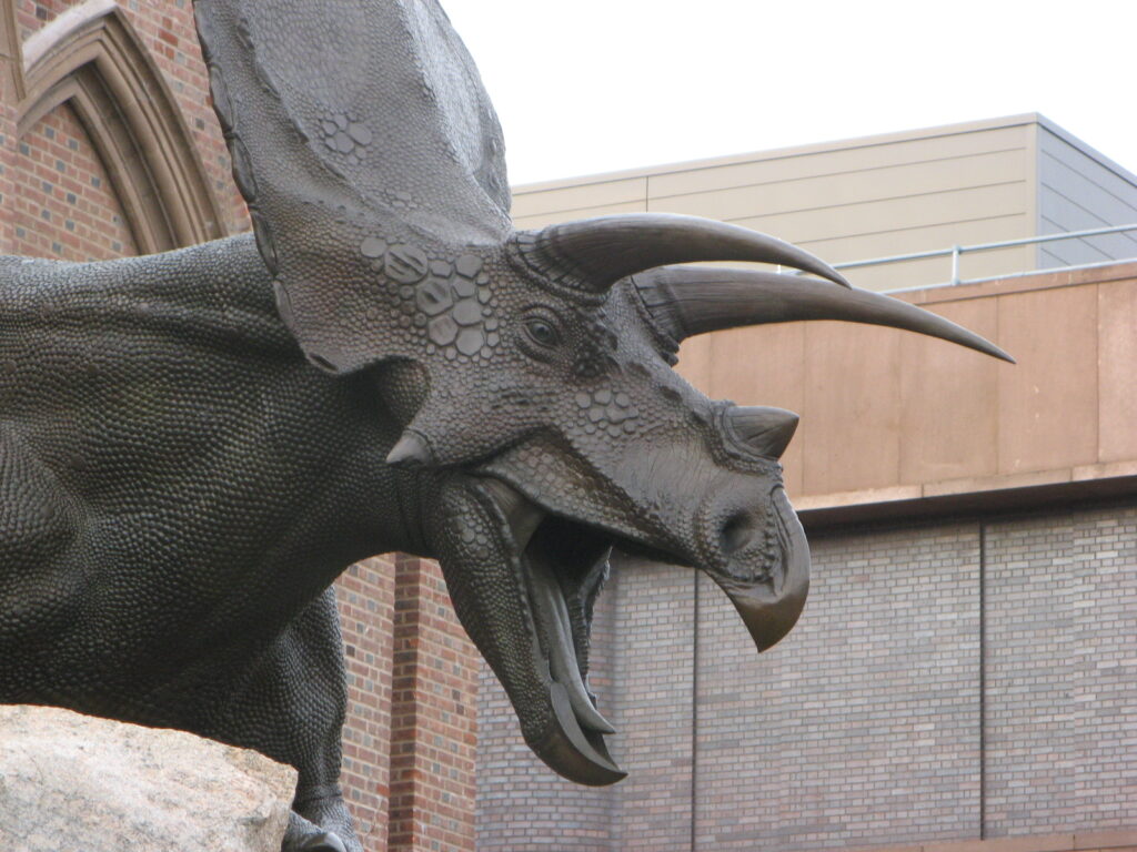 Sculpture of a horned dinosaur, possibly a torosaurus, with detailed skin texture and an open mouth, set against a backdrop of brick and modern buildings.