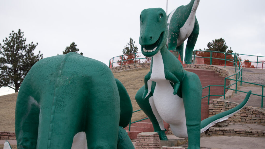 Two large green dinosaur sculptures stand in a park with surrounding trees, featuring bright expressions against a cloudy sky, creating a playful atmosphere.