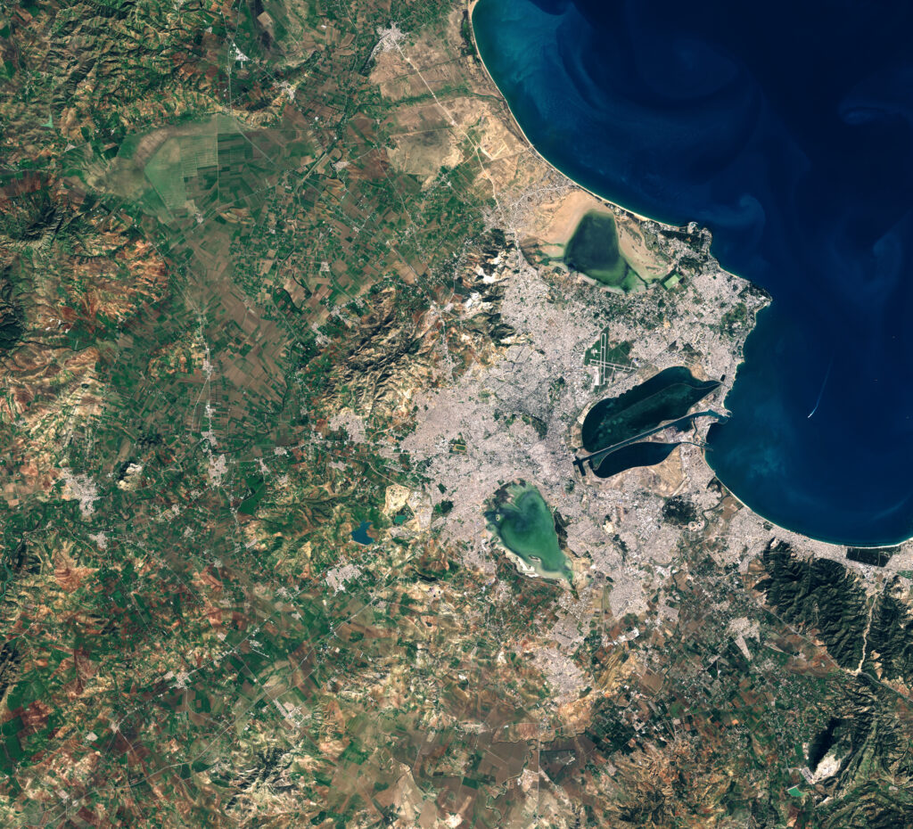 Satellite view of a coastal city with dense urban areas, green fields, and rugged terrain. The deep blue sea curves around the land in the top right.