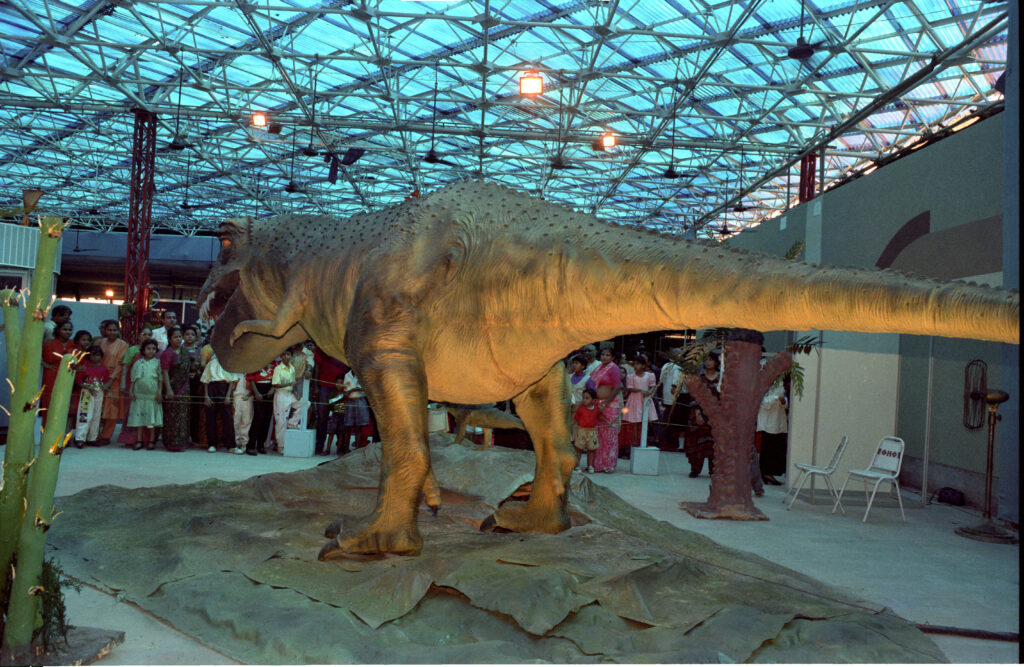 Lifelike dinosaur sculpture with textured skin displayed indoors. A crowd of people observes, set against a backdrop of geometric ceiling patterns.