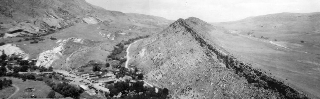 A black and white aerial view of a narrow, rocky ridge stretching diagonally across a vast, rural landscape. Small buildings and trees cluster in the foreground.
