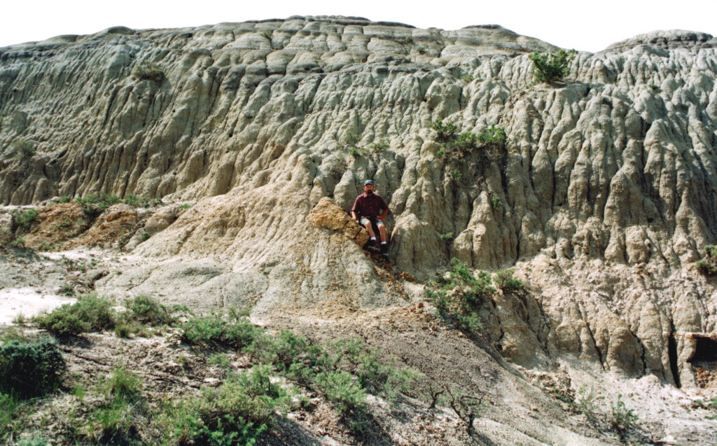 A person in a red shirt sits on rugged, eroded rock formations with vertical striations, surrounded by sparse greenery, conveying a sense of adventure.