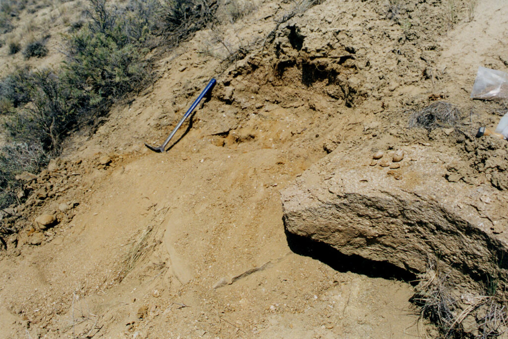 A rocky excavation site in an arid landscape with scattered shrubs. A geological hammer rests on loose earth beside rocks and a plastic bag.