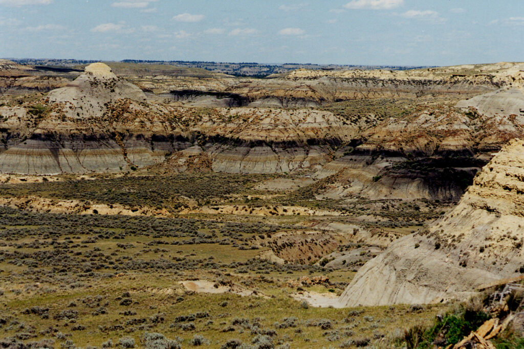 Arid landscape with layered rock formations in earthy tones, surrounded by sparse vegetation under a blue sky with scattered clouds. Rugged and expansive.