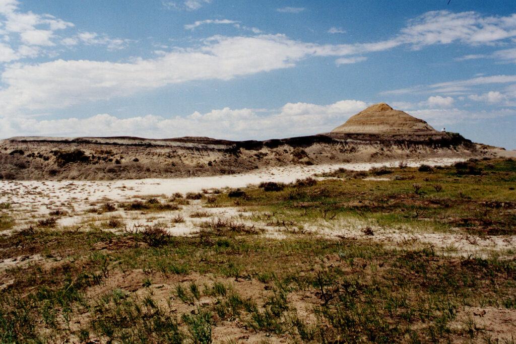 Arid landscape featuring a sunlit, rolling hill with stratified rock layers, sparse vegetation, and a broad, cloudy sky, conveying solitude and vastness.
