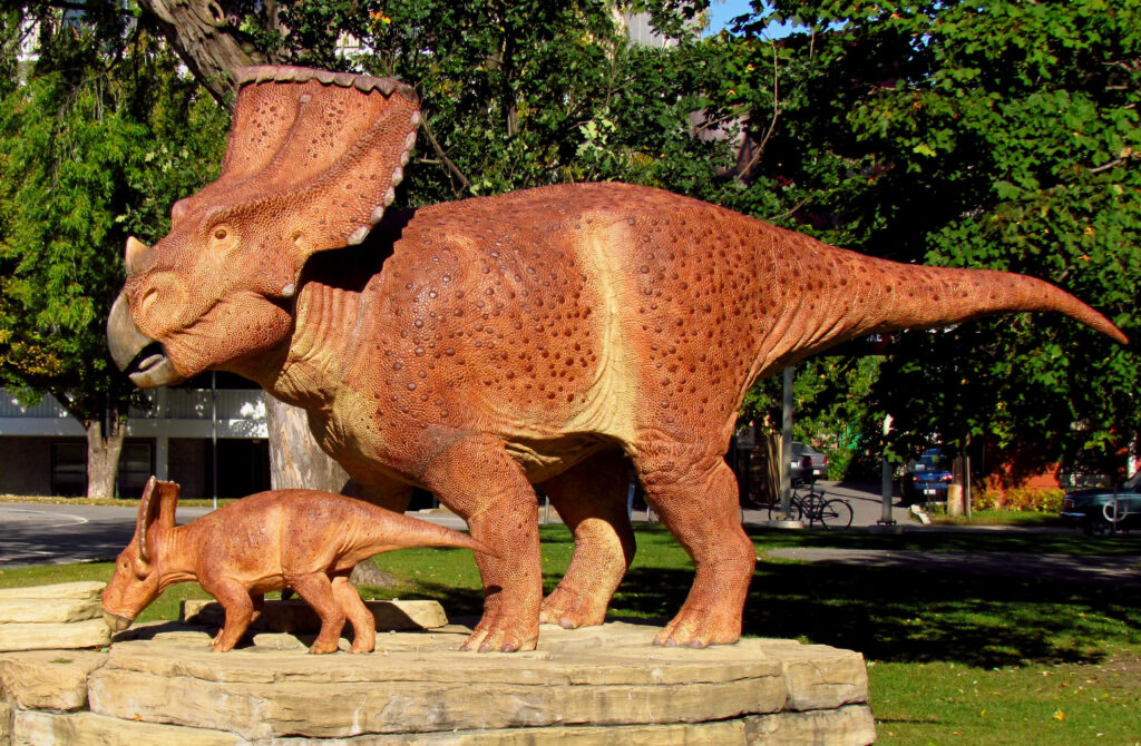 Statues of two brown horned dinosaurs, an adult and a juvenile, standing on rock slabs in a green, sunlit park setting.