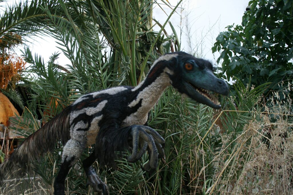 A realistic dinosaur sculpture, resembling a Velociraptor, stands amid lush greenery. The scene suggests an ancient, wild jungle environment.