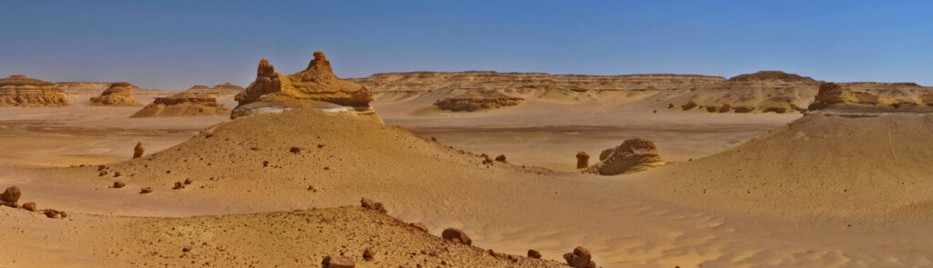Panoramic view of a vast desert landscape with rugged, eroded hills and scattered boulders under a clear blue sky. The scene feels expansive and desolate.