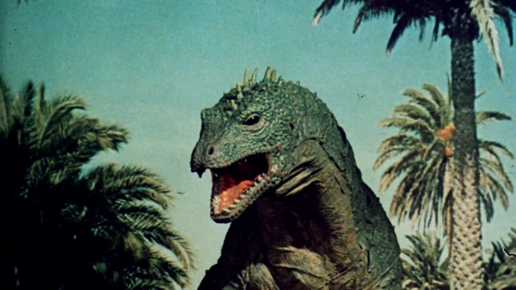 A large, green, scaly dinosaur with an open mouth stands amidst tall palm trees under a clear blue sky, conveying a sense of ancient, prehistoric life.
