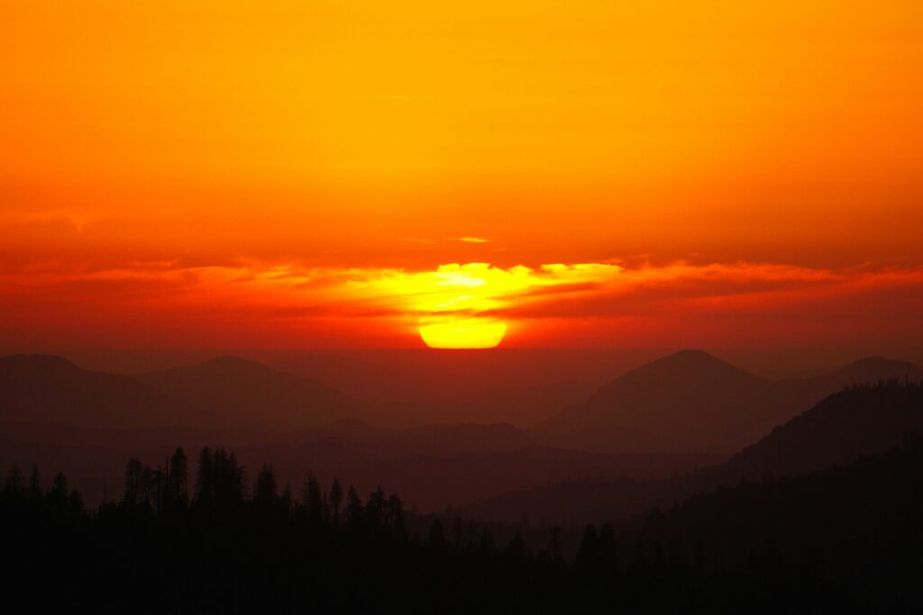 Vivid sunset over layered mountain silhouettes, with a fiery orange sky and the sun partially hidden by wispy clouds, creating a serene, warm atmosphere.