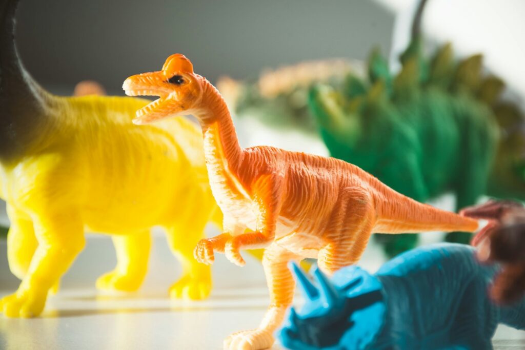 Toy dinosaurs in bright colors are displayed on a light surface. A prominent orange dinosaur is in focus, surrounded by yellow and green figures. Playful mood.