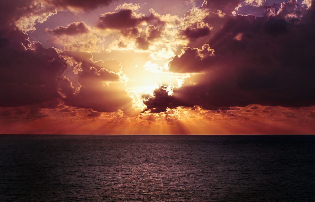 Dramatic sunset over the ocean with vibrant orange and purple clouds. Sunlight pierces through creating a warm, tranquil, and majestic atmosphere.