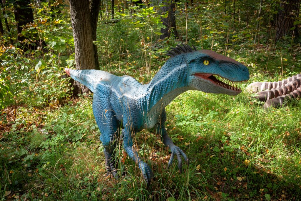 A blue dinosaur statue with yellow eyes and an open mouth stands on green grass in a dense forest, creating a lifelike and playful scene.