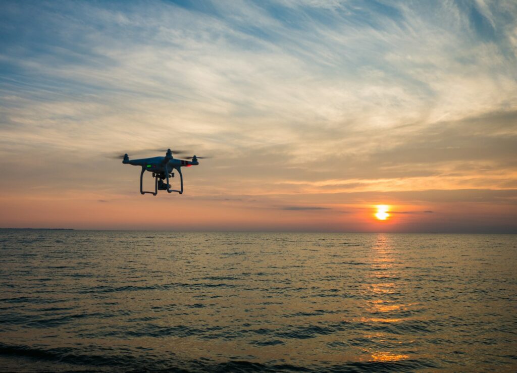 Drone hovering over a calm ocean during sunset. The sky is painted with soft pink and orange hues, creating a tranquil and serene atmosphere.
