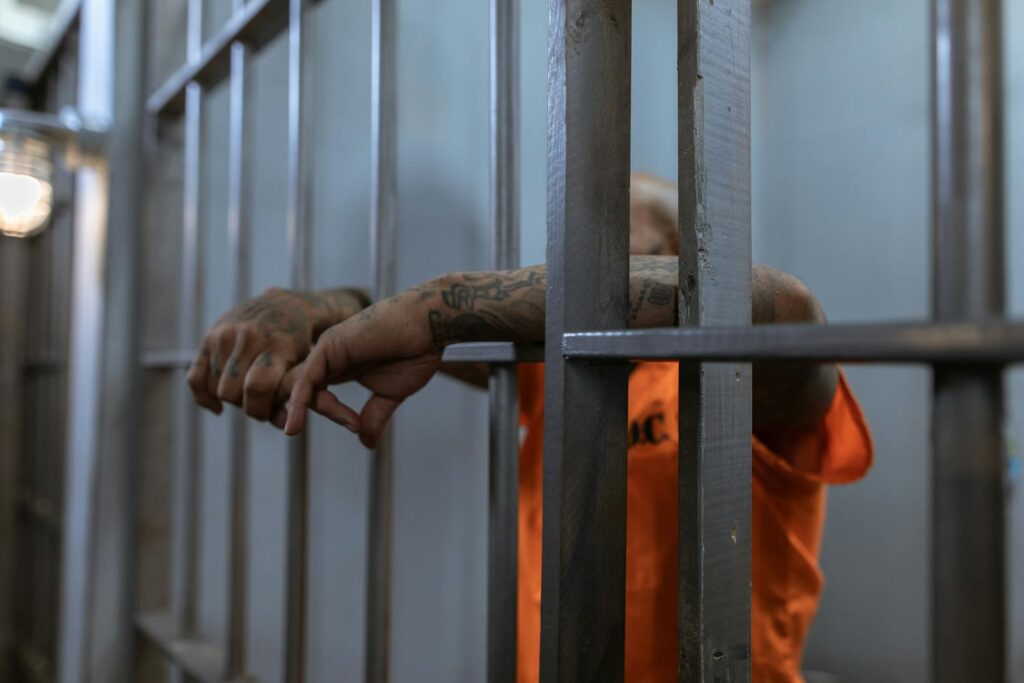 Tattooed arms in an orange jumpsuit rest on metal jail bars. The dim lighting conveys a somber mood, highlighting the confinement and isolation.