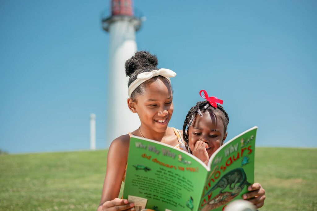 Two happy sisters reading a dinosaur book outdoors near a lighthouse.