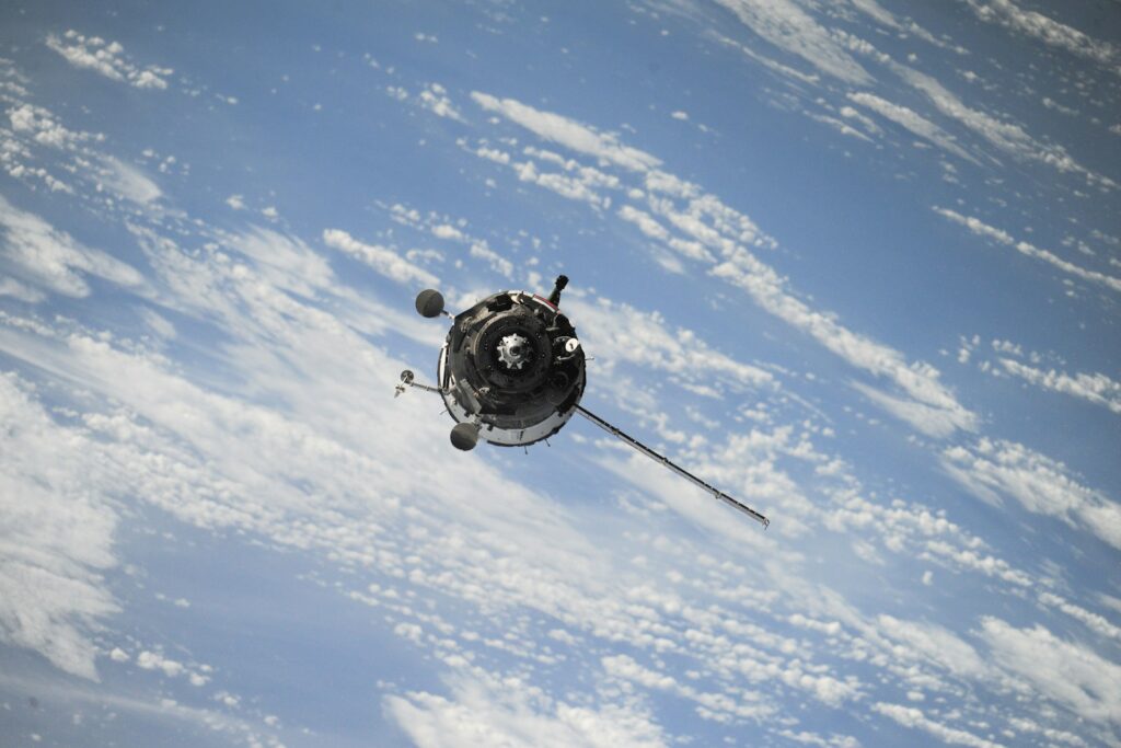A satellite orbits Earth against a backdrop of blue sky and scattered clouds. The scene conveys a sense of technological advancement and exploration.