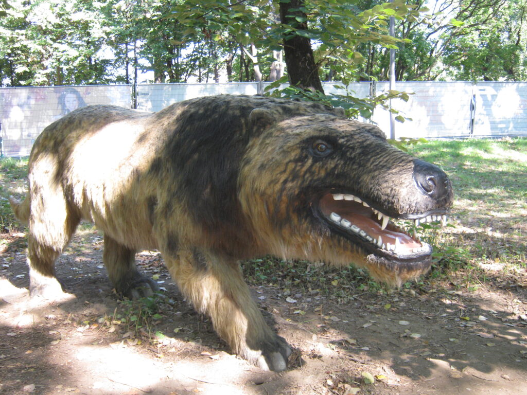 Lifelike sculpture of a prehistoric creature, resembling a large, furry wolf with sharp teeth, on a dirt path under dappled sunlight amidst trees.