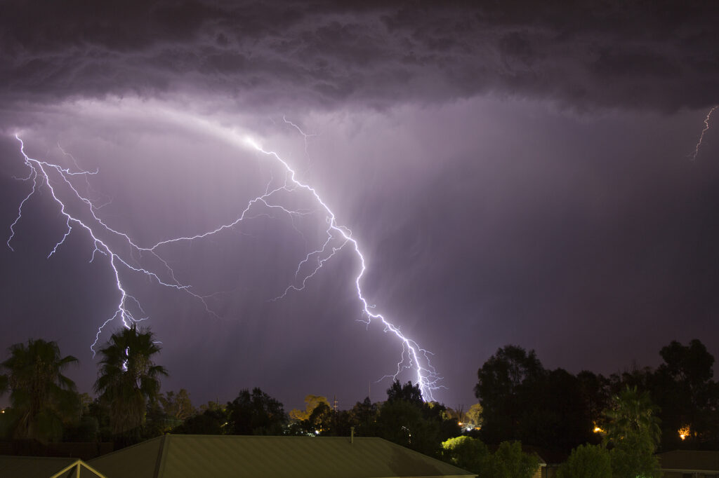 Dramatic nighttime scene of vivid lightning bolts illuminating a dark sky over silhouetted trees and rooftops, conveying a sense of power and intensity.