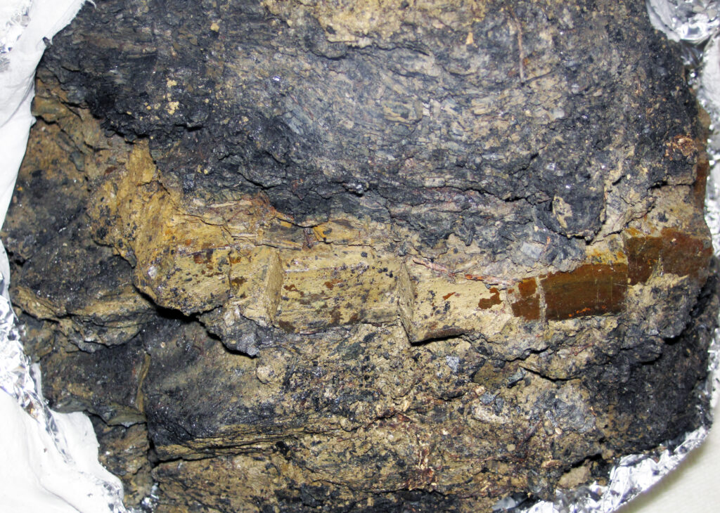 Close-up view of a rough, textured mineral rock surface with earthy brown and dark gray layers. Metallic sheen and embedded crystals add depth. Rugged and natural.