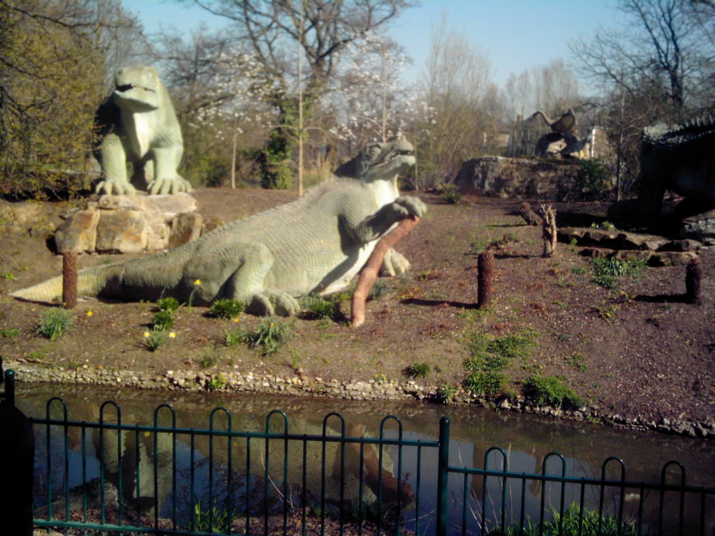 Large, green dinosaur sculptures sit in a park setting, surrounded by bare trees and a small pond. The scene evokes a calm, prehistoric atmosphere.