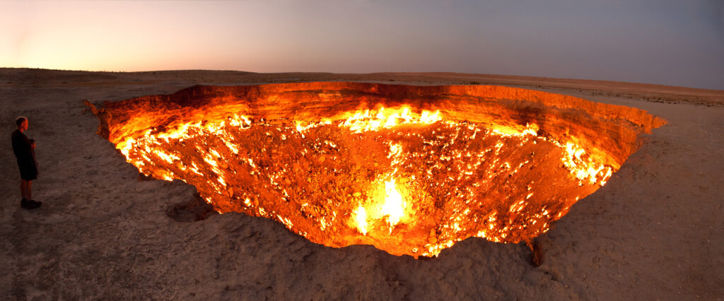 Person stands near the glowing, fiery pit of the Darvaza Gas Crater, known as the "Door to Hell," in a desert landscape at dusk, evoking awe.