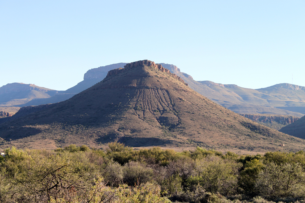 A sunlit, rugged mountain dominates the landscape with clear blue skies above and sparse green vegetation in the foreground, conveying a serene, open atmosphere.