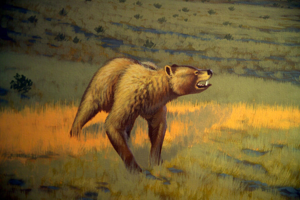 A saber-toothed, bear-like creature stands in a grassy landscape with a hunched posture and an open mouth showing sharp teeth. The tone is tense and dramatic.