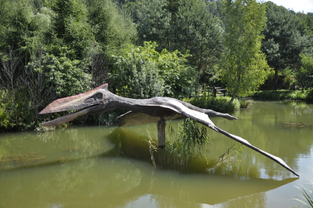 Quetzalcoatlus statue with open mouth and outstretched wings over a green pond, surrounded by lush green trees, conveying a prehistoric atmosphere.