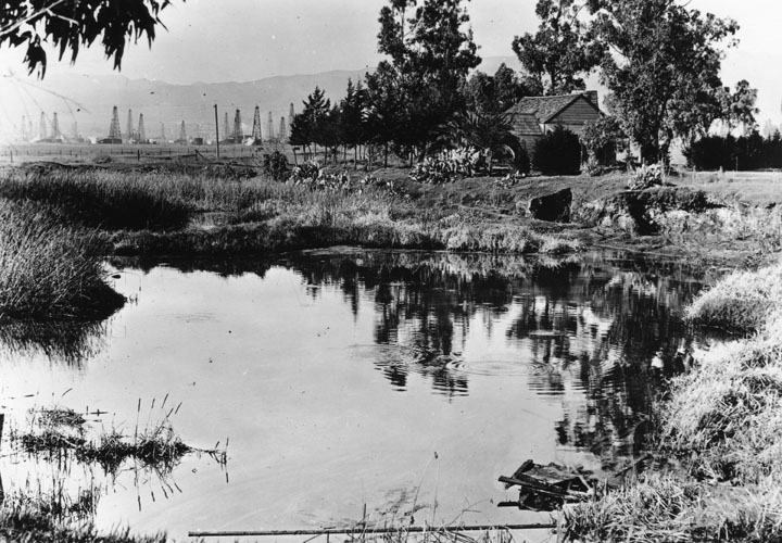 A serene pond reflecting trees and a rustic cabin in the foreground, with distant oil derricks and mountains under a hazy sky, conveying a tranquil rural-industrial contrast.
