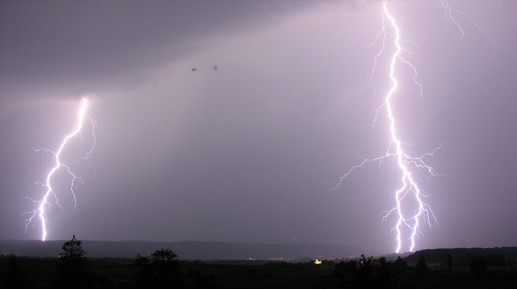 Lightning bolts illuminate a dark sky, striking the landscape below. Silhouettes of birds are visible near the powerful, bright flashes.