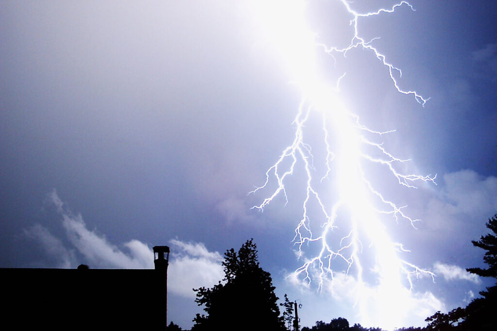 Dramatic lightning bolt illuminates a dark sky above silhouetted trees and a building. Intense and powerful, the scene conveys awe and energy.