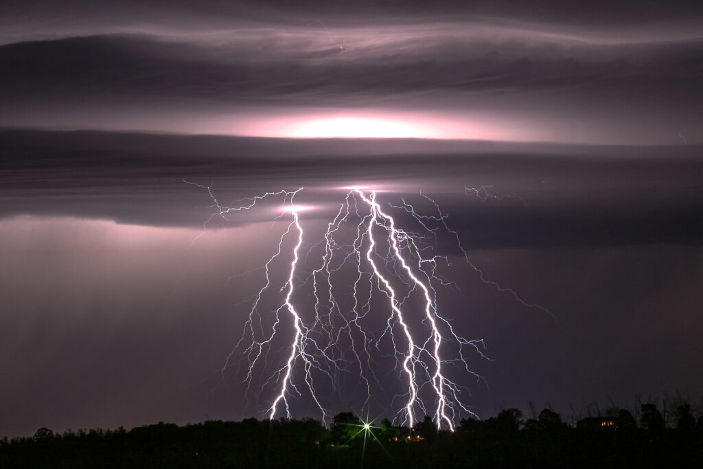 Dramatic lightning bolts illuminate the dark sky, striking silhouetted trees and a house on the horizon, creating a powerful and intense atmosphere.