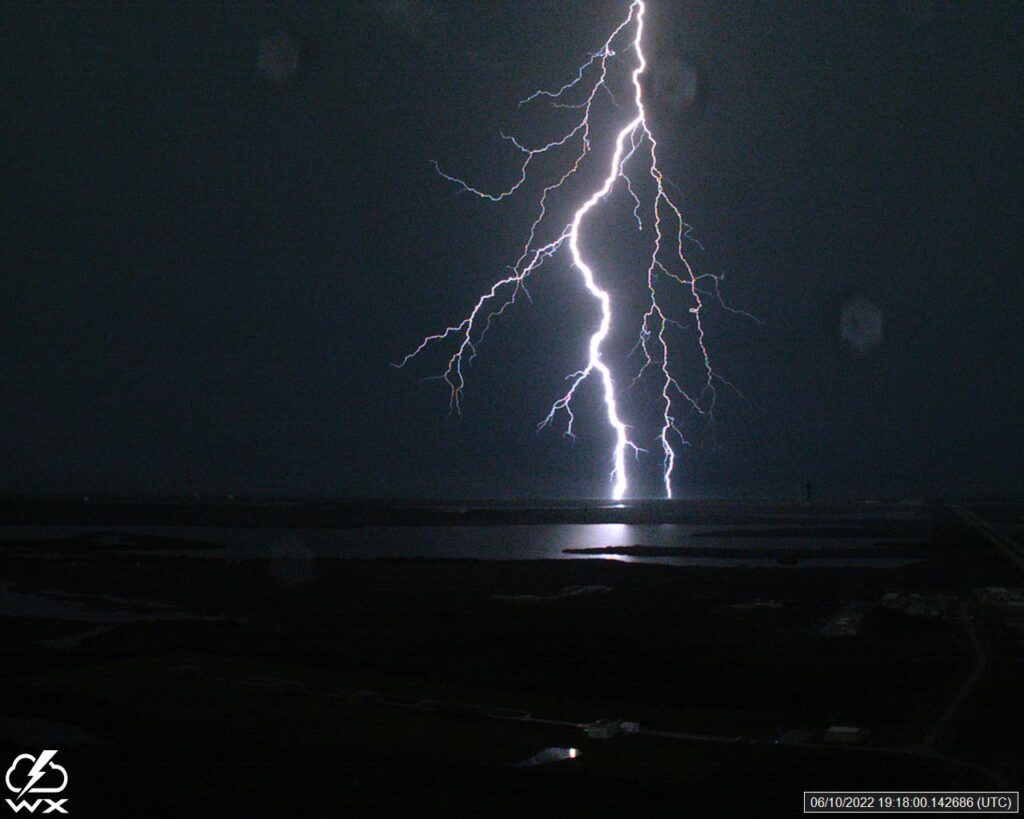 A bright lightning bolt illuminates a dark night sky, striking over a distant body of water. The scene conveys intensity and natural power.