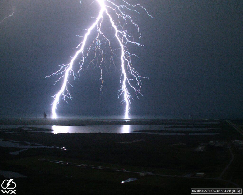 Intense lightning strikes over a dark coastal landscape, illuminating the night sky. Reflections shimmer on calm water, creating a dramatic, powerful scene.